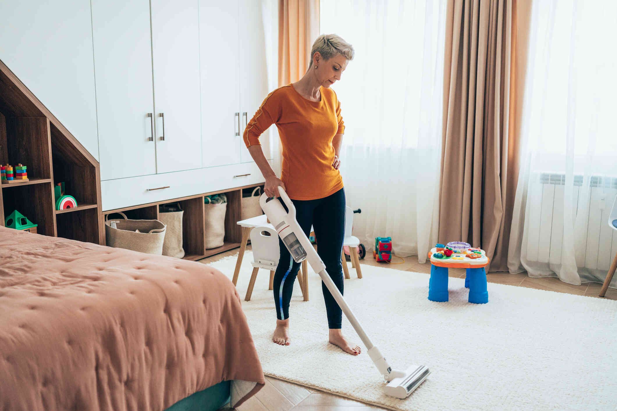 A woman in an orange shirt vacuums her living room with a focused expression.